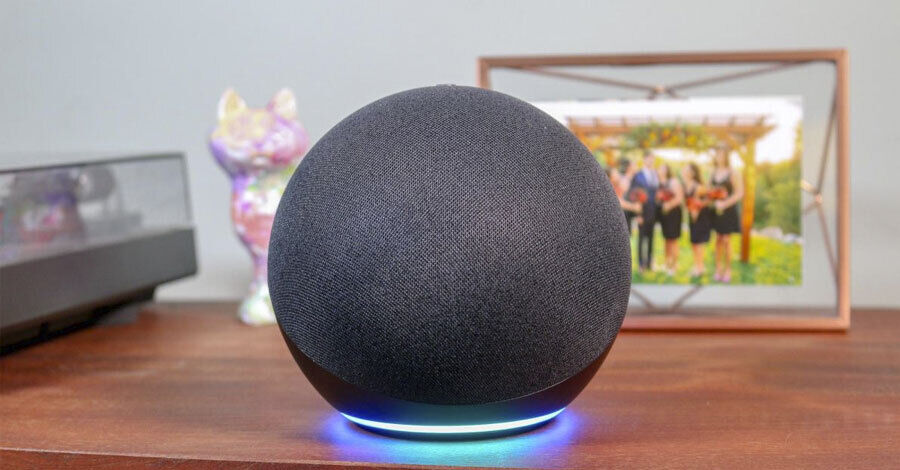 un dispositivo Amazon Alexa sobre una mesa, detrás se ve un portaretratos y adornos